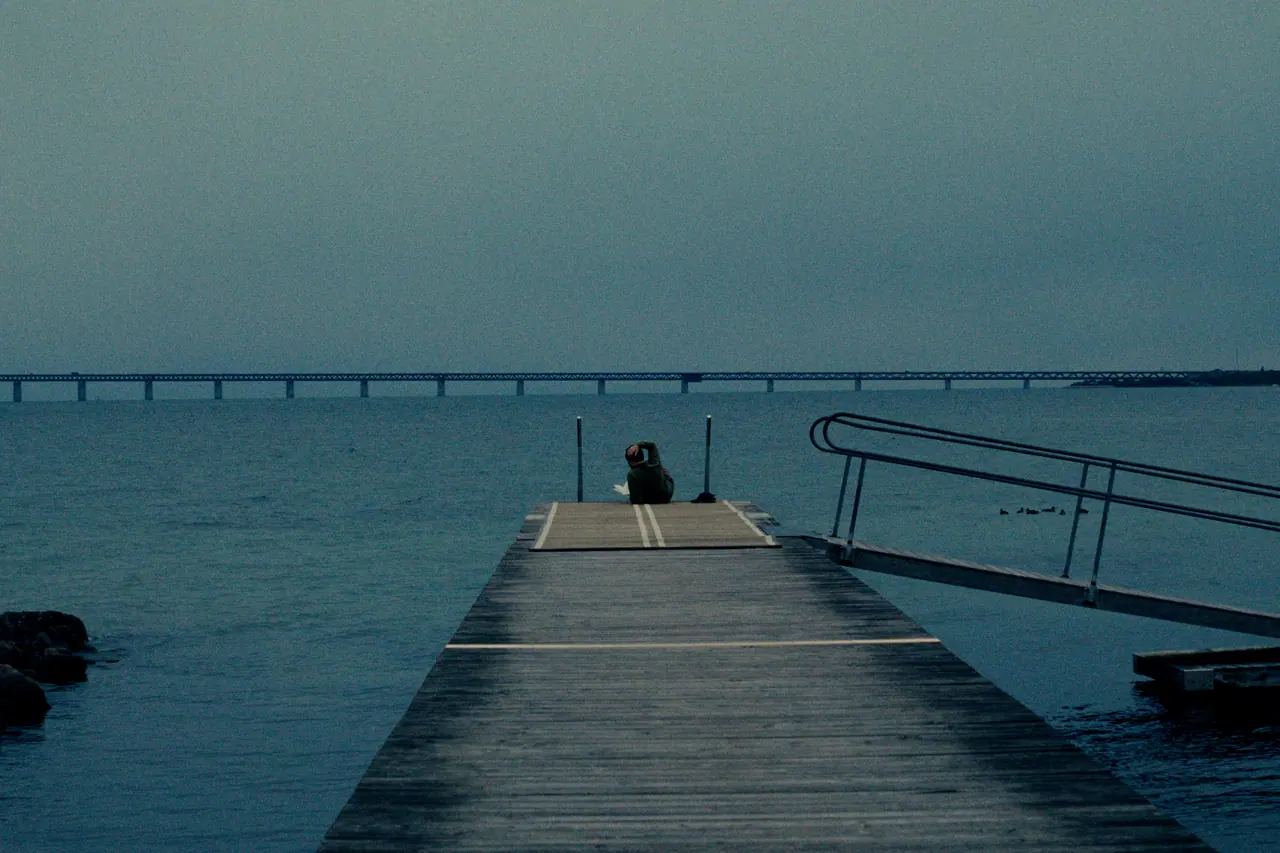 A lone person sitting at the end of a wooden pier at dusk, a long bridge on the horizon