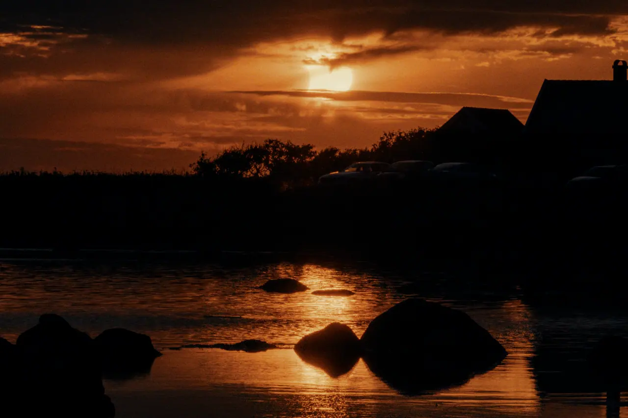 The sun breaking through clouds over a rocky shoreline, houses in silhouette